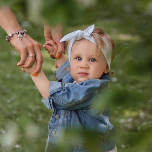 Cute baby with a headband holding hands, exploring outdoors in lush greenery.
