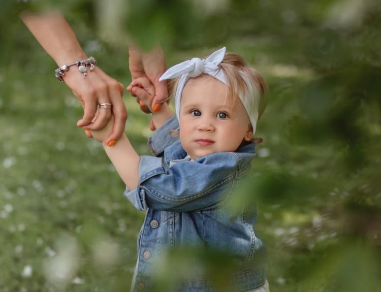 Cute baby with a headband holding hands, exploring outdoors in lush greenery.