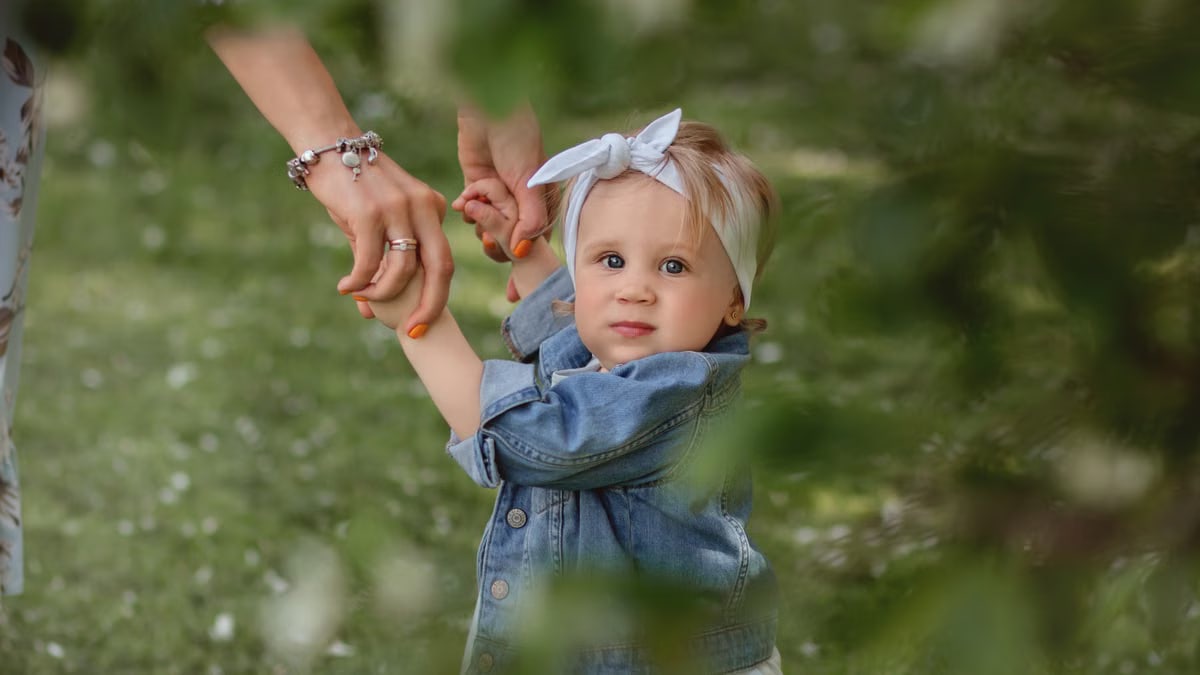Cute baby with a headband holding hands, exploring outdoors in lush greenery.